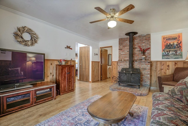 living room with a wood stove, wooden walls, ceiling fan, light wood-type flooring, and ornamental molding