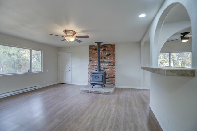 unfurnished living room featuring a wood stove, ceiling fan, a baseboard heating unit, and hardwood / wood-style flooring