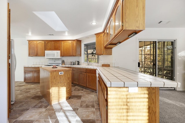 kitchen with a skylight, tile counters, a center island, kitchen peninsula, and white appliances