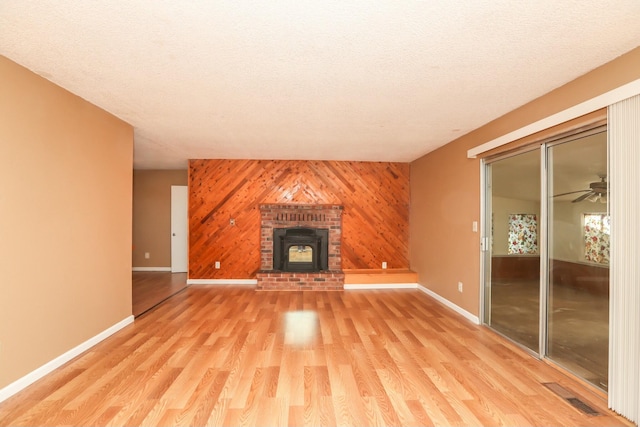 unfurnished living room featuring a wood stove, ceiling fan, a textured ceiling, wooden walls, and light wood-type flooring