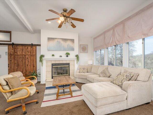 living room featuring dark carpet, a fireplace, ceiling fan, a barn door, and beamed ceiling