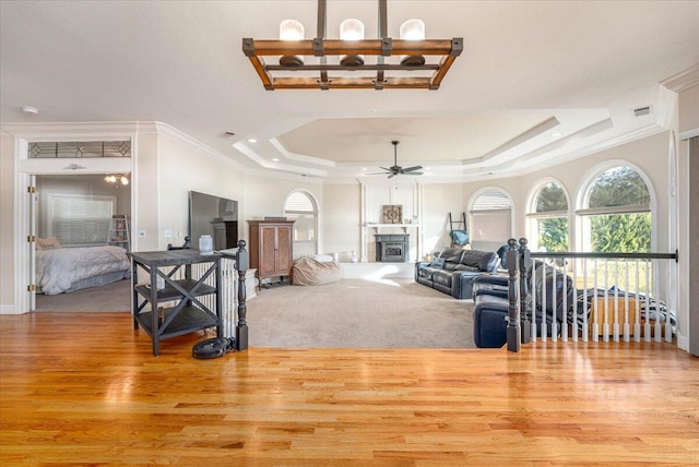 living room with light wood-type flooring, ornamental molding, ceiling fan with notable chandelier, and a tray ceiling