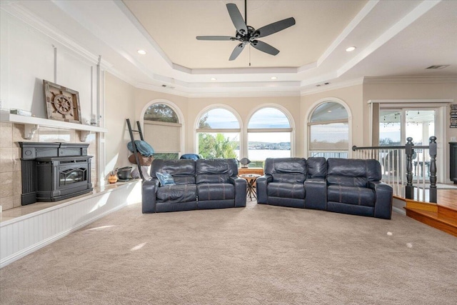 living room featuring ceiling fan, a wood stove, carpet flooring, a raised ceiling, and ornamental molding