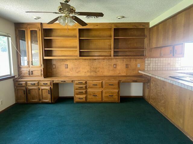 unfurnished office featuring ceiling fan, built in desk, a textured ceiling, and dark colored carpet