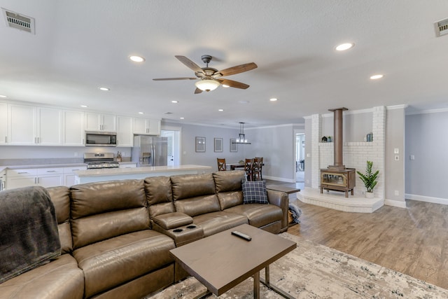 living room featuring light wood-type flooring, ceiling fan, ornamental molding, and a wood stove