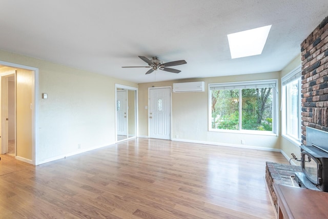 unfurnished living room featuring a wood stove, ceiling fan, light hardwood / wood-style floors, a skylight, and a wall mounted air conditioner