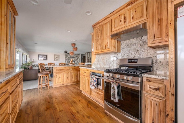 kitchen featuring a kitchen bar, decorative backsplash, stainless steel range with gas stovetop, light wood-type flooring, and light stone counters