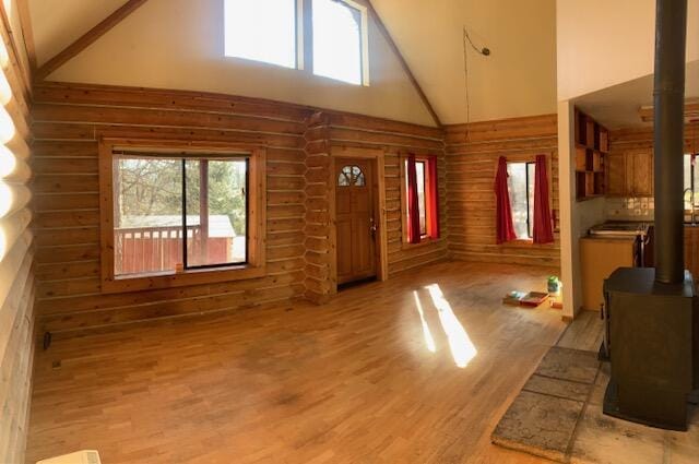 unfurnished living room featuring plenty of natural light, high vaulted ceiling, light hardwood / wood-style floors, log walls, and a wood stove