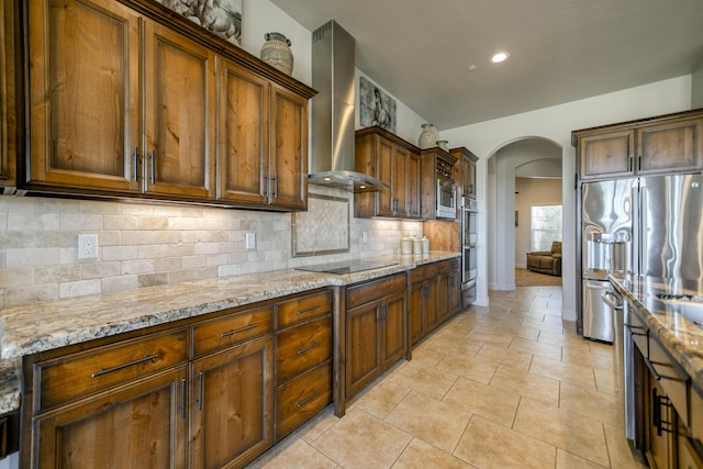 kitchen featuring light tile patterned floors, appliances with stainless steel finishes, light stone counters, decorative backsplash, and wall chimney exhaust hood