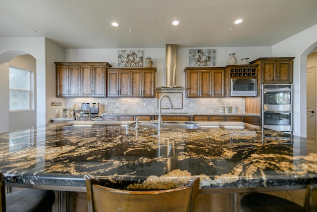 kitchen with wall chimney exhaust hood, stainless steel appliances, sink, and dark stone counters