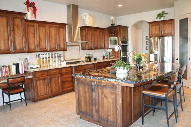 kitchen with appliances with stainless steel finishes, dark stone counters, a kitchen island with sink, decorative backsplash, and exhaust hood