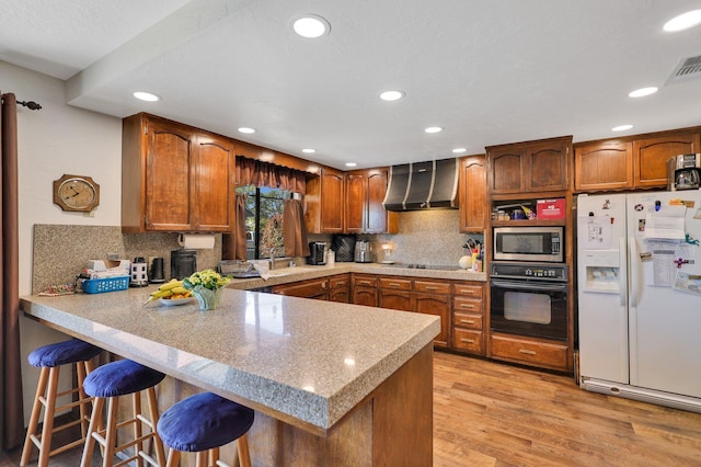 kitchen with wall chimney range hood, light hardwood / wood-style flooring, black appliances, a kitchen bar, and kitchen peninsula