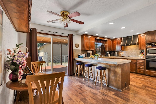 kitchen with wall chimney exhaust hood, a kitchen bar, kitchen peninsula, oven, and hardwood / wood-style floors