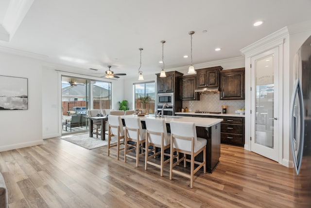 kitchen with dark brown cabinets, hanging light fixtures, stainless steel appliances, a kitchen island with sink, and decorative backsplash