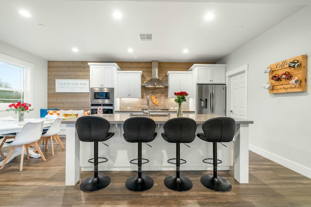 kitchen with white cabinetry, wall chimney range hood, a kitchen island with sink, and appliances with stainless steel finishes