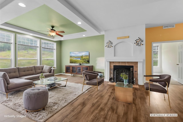 living area with a tiled fireplace, visible vents, a tray ceiling, and wood finished floors