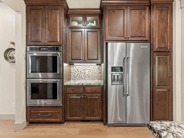 kitchen with appliances with stainless steel finishes, dark brown cabinets, light stone countertops, decorative backsplash, and light wood-type flooring