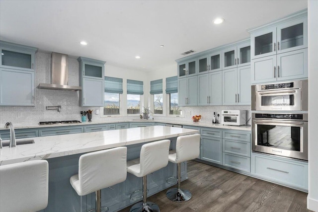 kitchen with wall chimney exhaust hood, light stone counters, dark hardwood / wood-style floors, stainless steel appliances, and backsplash