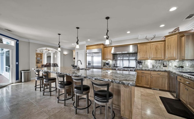 kitchen featuring hanging light fixtures, an island with sink, stainless steel appliances, and wall chimney exhaust hood