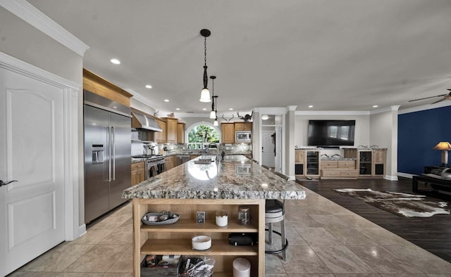 kitchen featuring an island with sink, decorative light fixtures, built in appliances, extractor fan, and a kitchen breakfast bar