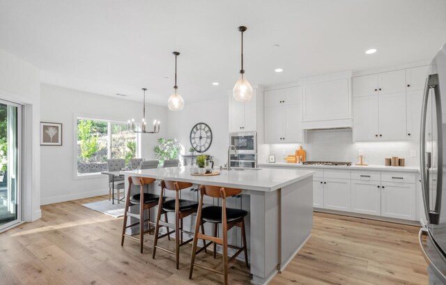 kitchen with white cabinetry, light countertops, appliances with stainless steel finishes, an island with sink, and decorative light fixtures