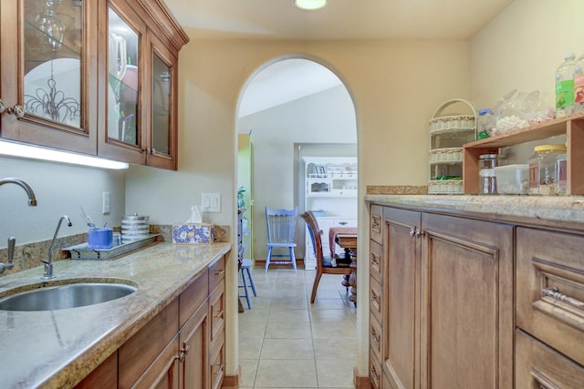 kitchen featuring arched walkways, light stone counters, light tile patterned flooring, a sink, and glass insert cabinets