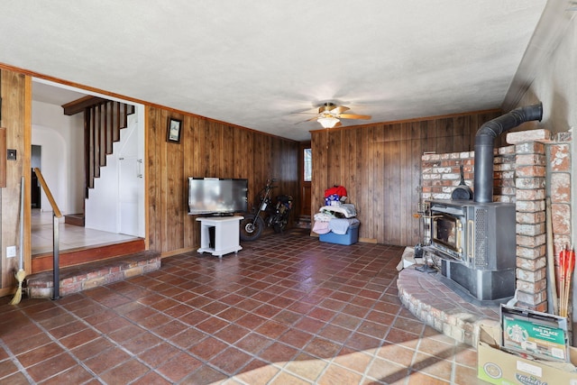 unfurnished living room featuring a wood stove, a ceiling fan, wood walls, tile patterned flooring, and stairs