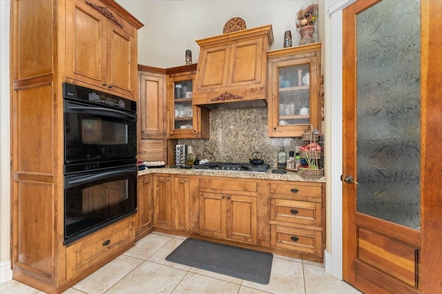 kitchen with brown cabinets, light tile patterned floors, tasteful backsplash, dobule oven black, and glass insert cabinets