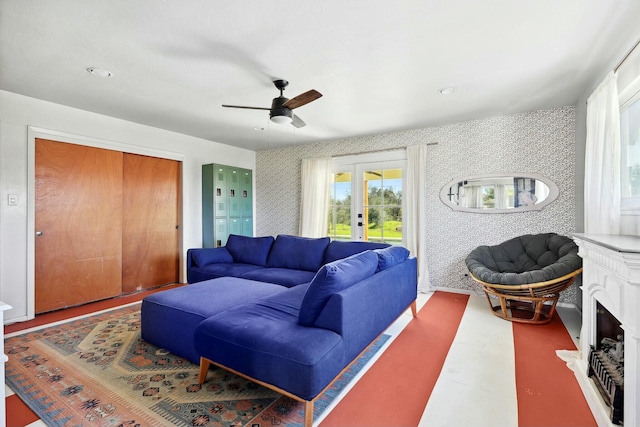 living room featuring wallpapered walls, ceiling fan, a fireplace with raised hearth, and french doors