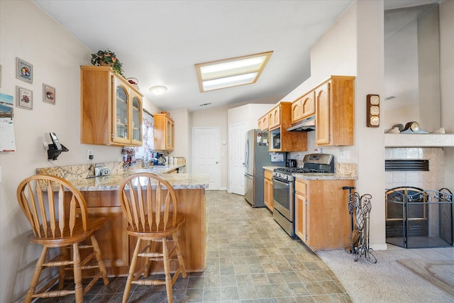 kitchen with light stone counters, appliances with stainless steel finishes, glass insert cabinets, a peninsula, and under cabinet range hood