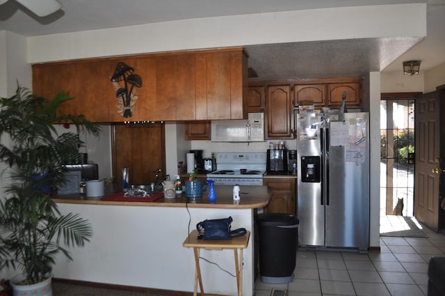 kitchen featuring light tile patterned floors, a textured ceiling, a peninsula, white appliances, and brown cabinets