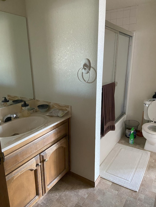 bathroom with toilet, stone finish floor, bath / shower combo with glass door, and vanity