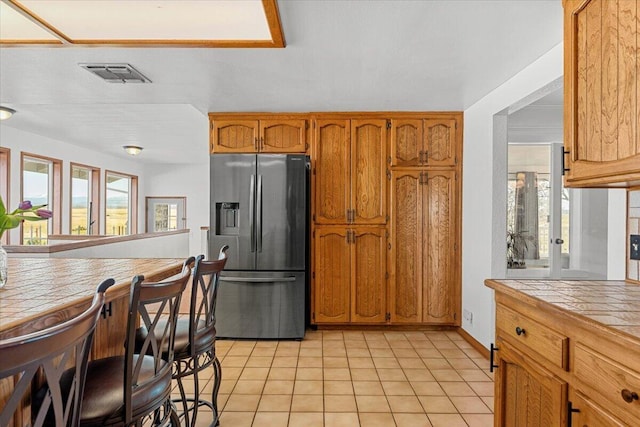 kitchen featuring light tile patterned floors, visible vents, tile countertops, brown cabinets, and stainless steel refrigerator with ice dispenser