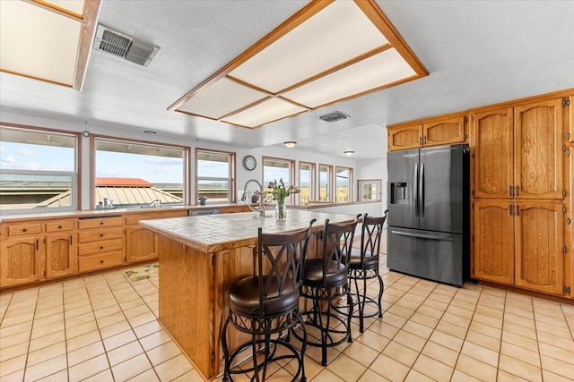 kitchen featuring light tile patterned floors, visible vents, an island with sink, stainless steel fridge, and a kitchen bar