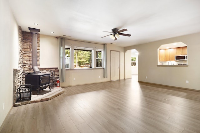 unfurnished living room with baseboards, arched walkways, ceiling fan, wood finished floors, and a wood stove