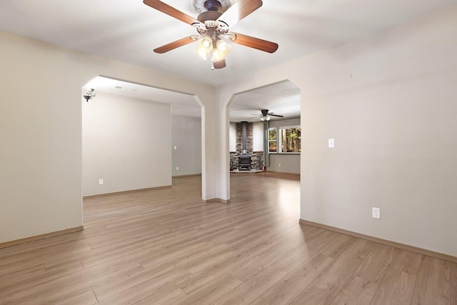 unfurnished room featuring arched walkways, a wood stove, ceiling fan, light wood-type flooring, and baseboards