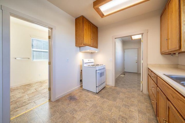 kitchen featuring under cabinet range hood, brown cabinetry, light countertops, baseboards, and white gas range