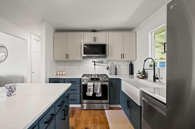 kitchen with backsplash, blue cabinetry, light wood-type flooring, appliances with stainless steel finishes, and a sink