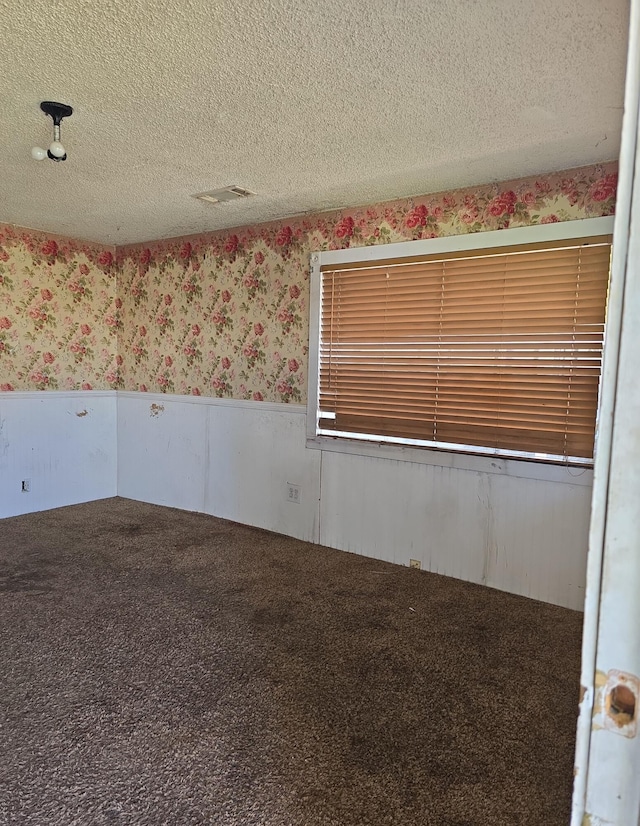 carpeted empty room with visible vents, a textured ceiling, wainscoting, and wallpapered walls