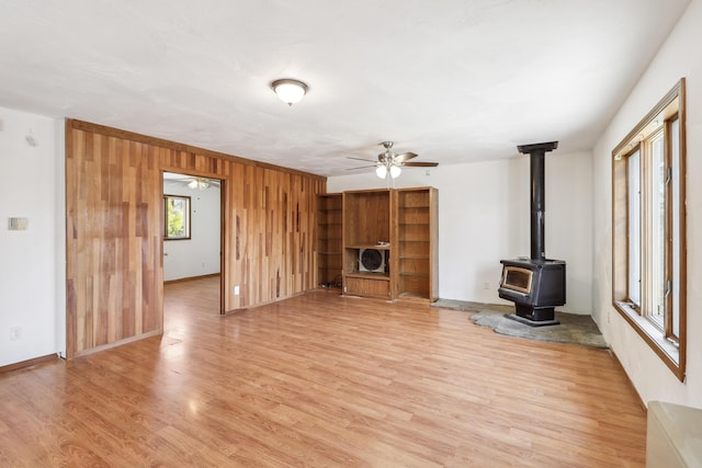 unfurnished living room with a ceiling fan, baseboards, light wood finished floors, a wood stove, and wood walls