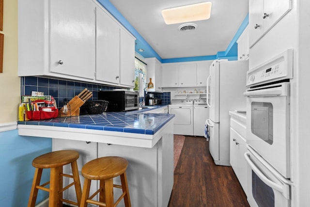 kitchen with tile countertops, white appliances, independent washer and dryer, a peninsula, and white cabinets