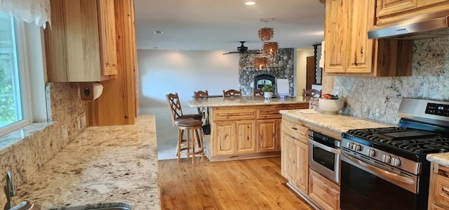 kitchen featuring stainless steel range with gas stovetop, under cabinet range hood, a kitchen bar, ceiling fan, and light wood-style flooring