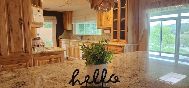 kitchen with decorative backsplash, glass insert cabinets, a sink, light stone countertops, and a ceiling fan