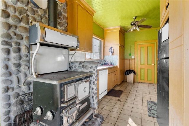kitchen featuring light tile patterned floors, a ceiling fan, a wainscoted wall, a wood stove, and white dishwasher