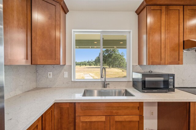 kitchen with a sink, brown cabinetry, backsplash, light stone counters, and black microwave