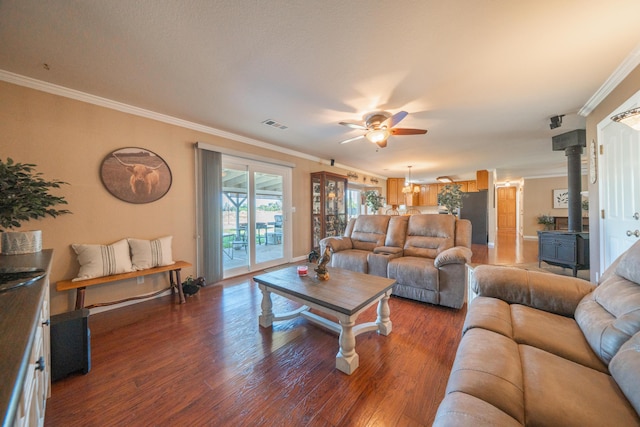 living area with wood finished floors, visible vents, ceiling fan, a wood stove, and ornamental molding