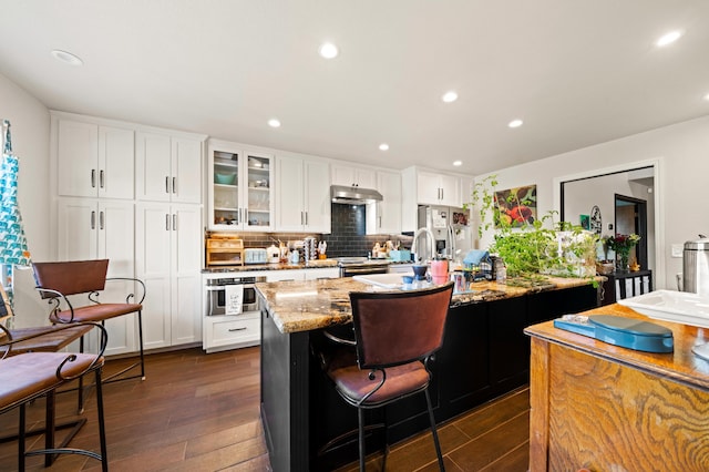 kitchen with a kitchen island with sink, stainless steel appliances, dark wood finished floors, light stone countertops, and under cabinet range hood