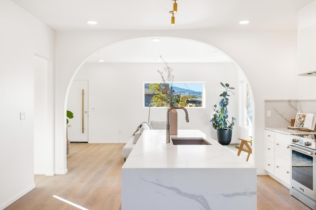 kitchen with arched walkways, a sink, a center island with sink, stainless steel electric range oven, and white cabinetry