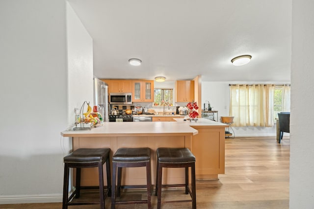 kitchen with light brown cabinets, light wood-type flooring, baseboards, a peninsula, and appliances with stainless steel finishes