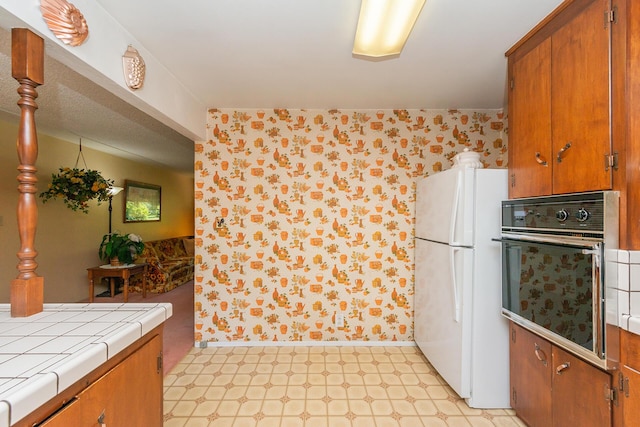 kitchen with tile counters, light floors, freestanding refrigerator, wall oven, and brown cabinetry